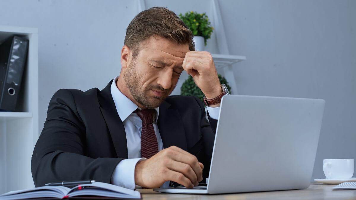 Muškarac u uredu drži se za čelo dok radi na laptopu, simbol glavobolje nakon kave i napetosti tijekom radnog dana.